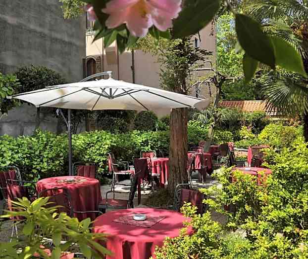 En frodig gårdsplass med røde bord og stoler samt en stor hvit parasoll. (AI-generert tekst)