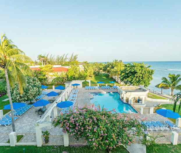 Bilde av et hotellområde ved sjøen med basseng, solsenger og parasoller omgitt av grøntområder og palmer. (AI-generert tekst)