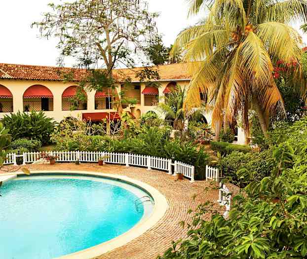 Et rundt svømmebasseng omgitt av et steinbelagt område og hvite gjerder. I bakgrunnen er en bygning med buede vinduer og røde markiser samt grønne omkringliggende planter. (AI-generert tekst)