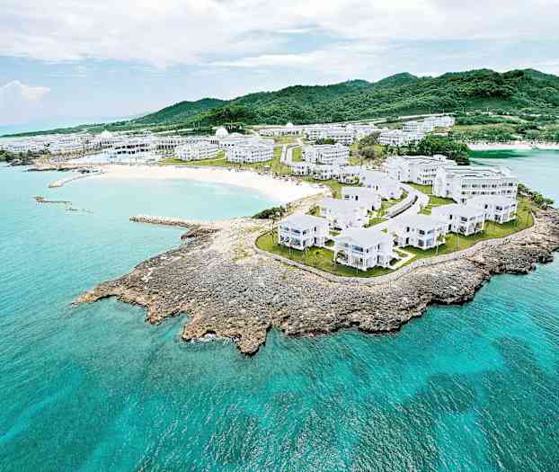 Luftfoto av et kystlandskap med hvite bygninger omgitt av grøntområder og turkist vann. (AI-generert tekst)