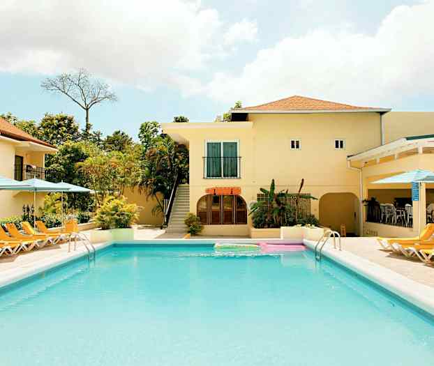 Et basseng med solsenger og parasoller rundt. Bak bassenget ligger en bygning med lyse vegger og et tak med terrakottafarget takstein. (AI-generert tekst)