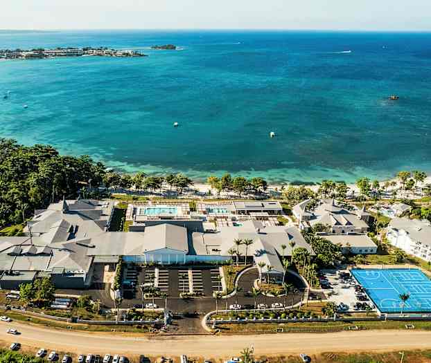 Luftfoto av et kystresort med bassenger, bygninger og en tennisbane ved havet. (AI-generert tekst)