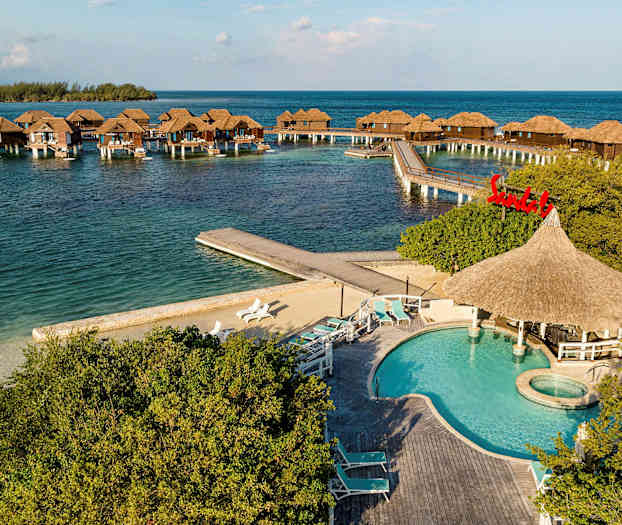 Flytende bungalower på stolper i havet med et basseng og solsenger i forgrunnen. (AI-generert tekst)