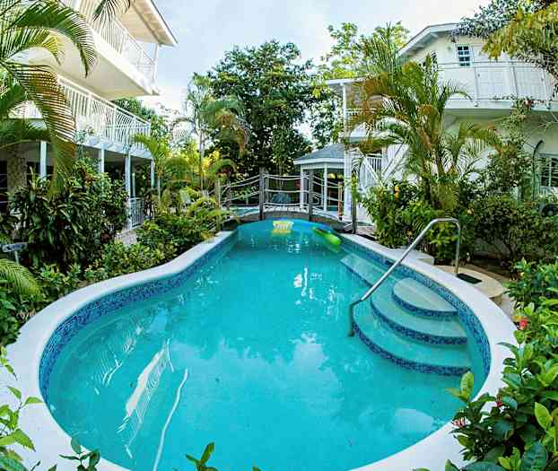 Et utendørsbasseng omgitt av grønne planter med bygninger i bakgrunnen. (AI-generert tekst)