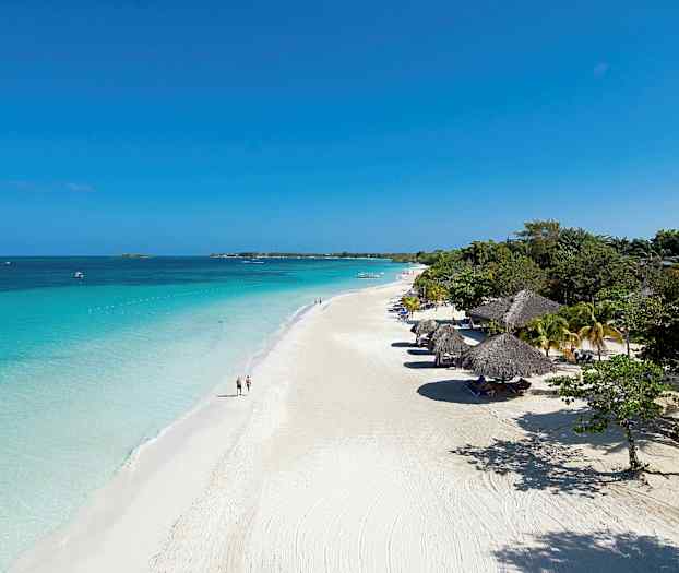 En hvit sandstrand med parasolltak og klart blått vann, omgitt av grøntområder. (AI-generert tekst)