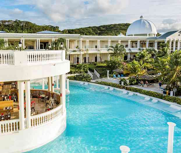 En stor bygning med hvite søyler og kuppel ved et basseng omgitt av palmer og solsenger. (AI-generert tekst)