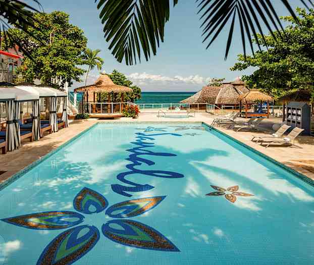 Et basseng med solsenger rundt og en bygning med stråtak synlig ved havet. (AI-generert tekst)