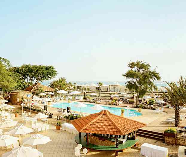 Bilde av et utendørsområde ved et basseng med parasoller og trær under klar himmel. (AI-generert tekst)