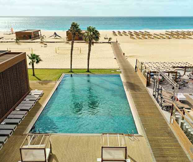 Utendørsbasseng med solsenger, plen, strand med parasoller og havet i bakgrunnen. (AI-generert tekst)