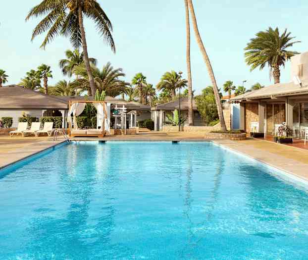 Et svømmebasseng omgitt av solsenger, en seng med gardiner og palmer i bakgrunnen. (AI-generert tekst)