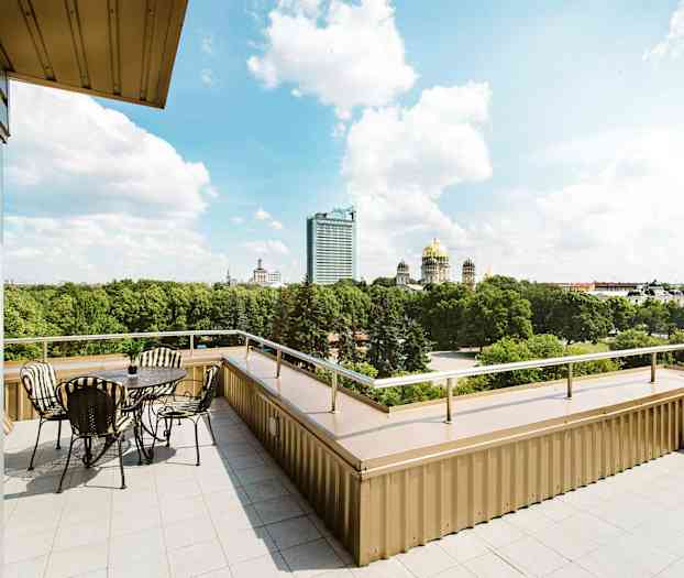 En terrasse med bord og stoler samt utsikt over en park og flere bygninger, inkludert en med gullkuppel. (AI-generert tekst)