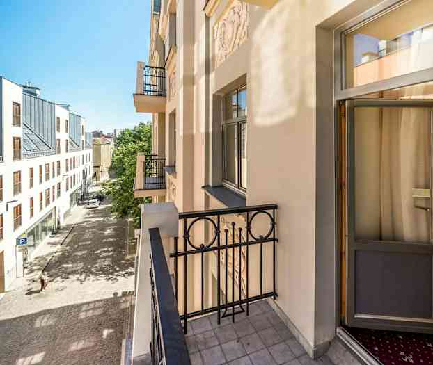 Bildet viser en balkong på en lys bygning med utsikt over en brosteinsgate og et moderne hus med store vinduer på den andre siden av gaten. (AI-generert tekst)