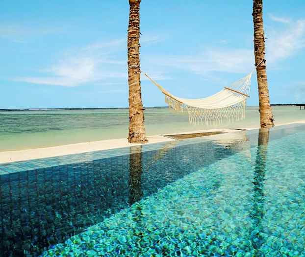 En hengekøye hengt mellom to palmer ved et basseng og en tropisk strand. (AI-generert tekst)