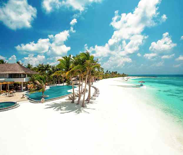 Bilde av en hvit sandstrand med turkis vann, solsenger, parasoller og palmetrær ved et hotellområde. (AI-generert tekst)