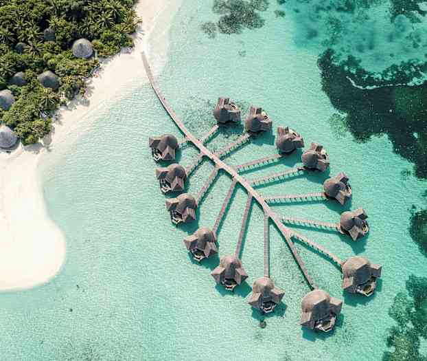 Flyfoto av bungalower på påler over klart blått vann med en nærliggende sandstrand og frodig vegetasjon. (AI-generert tekst)