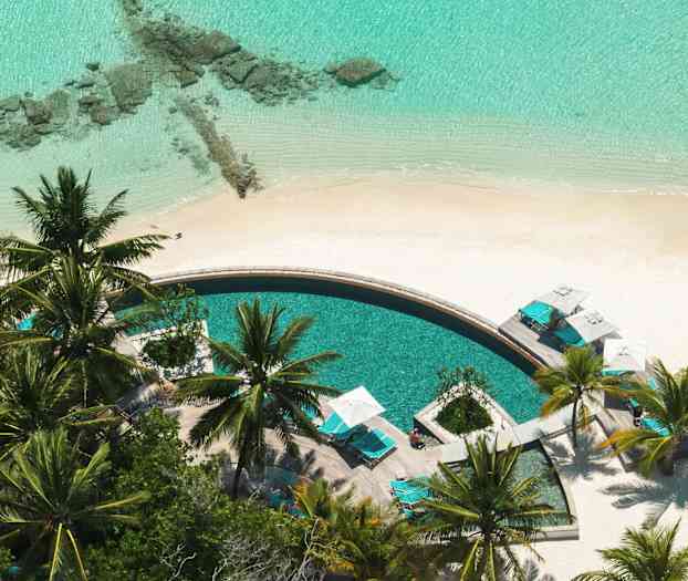 Et rundt basseng omgitt av solsenger og palmer ved en sandstrand. (AI-generert tekst)