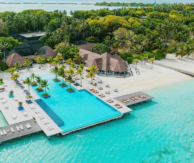 En resort med et basseng som ligger rett ved turkisblått vann, omgitt av palmer og grønt. (AI-generert tekst)