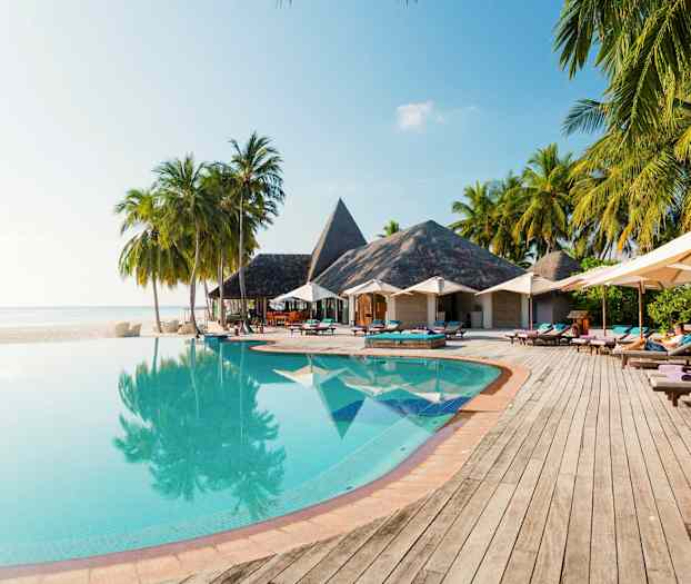 Et evighetsbasseng med trær og parasoller ved siden, samt bygninger med stråtak i bakgrunnen. (AI-generert tekst)