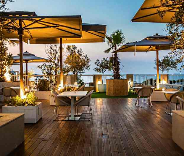 Bilde av en utendørsterrasse med tregulv, bord og stoler under parasoller, omgitt av grøntomgivelser i skumringslys. (AI-generert tekst)