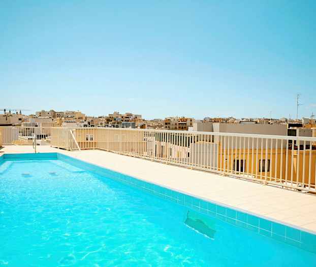 Et utendørs svømmebasseng med klart vann på en takterrasse. I bakgrunnen ses bybebyggelse under en klar blå himmel. (AI-generert tekst)