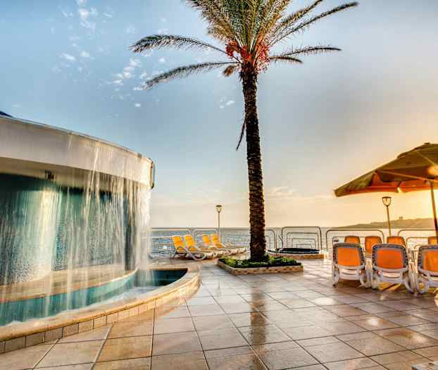 Et basseng med et fossefall ved siden av en palme, solsenger og utsikt over havet på en solrik dag. (AI-generert tekst)