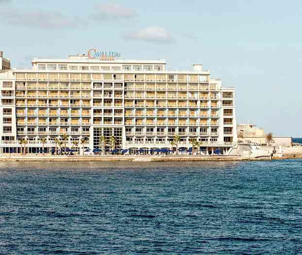 Et stort hotell ved sjøen, med mange etasjer og balkonger samt en liten båt i vannet nær hotellet. (AI-generert tekst)