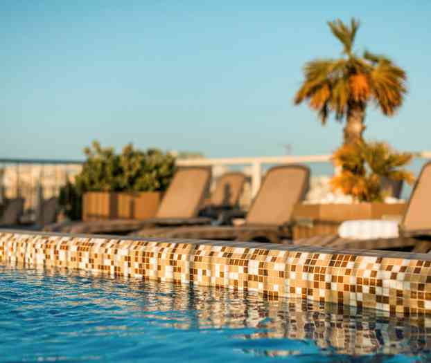 Nærbilde av en bassengkanten med klart blått vann og solsenger samt planter i bakgrunnen. (AI-generert tekst)