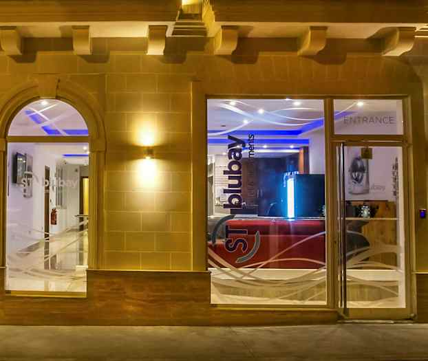 Inngangen til en bygning med glassvinduer og dører, opplyst om kvelden. (AI-generert tekst)