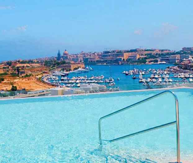 Utsikt over et basseng med rekkverk, hvor en havn med mange båter og bymiljø ses i bakgrunnen. (AI-generert tekst)