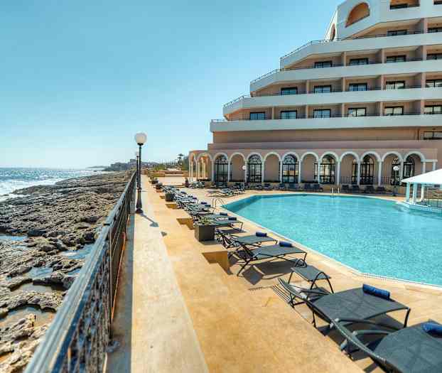 Bilde som viser et luksuriøst feriested ved sjøen med et svømmebasseng, solsenger og en bygning med balkonger. (AI-generert tekst)