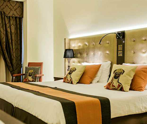 Et hotellrom med en stor oppredd seng, dekorert med puter med hundemotiv. Rommet har mørke gardiner og en stol med en matchende pute. (AI-generert tekst)