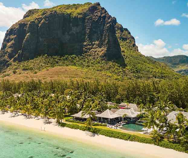 Et stort fjell vises i bakgrunnen av en strand med turkis vann i forgrunnen. På stranden står det palmetrær og bygninger med stråtak. (AI-generert tekst)