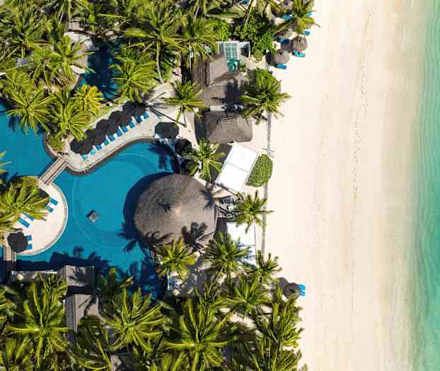 Bildet viser et luftperspektiv over en strand med hvit sand, turkist vann, og et basseng omgitt av palmer. (AI-generert tekst)