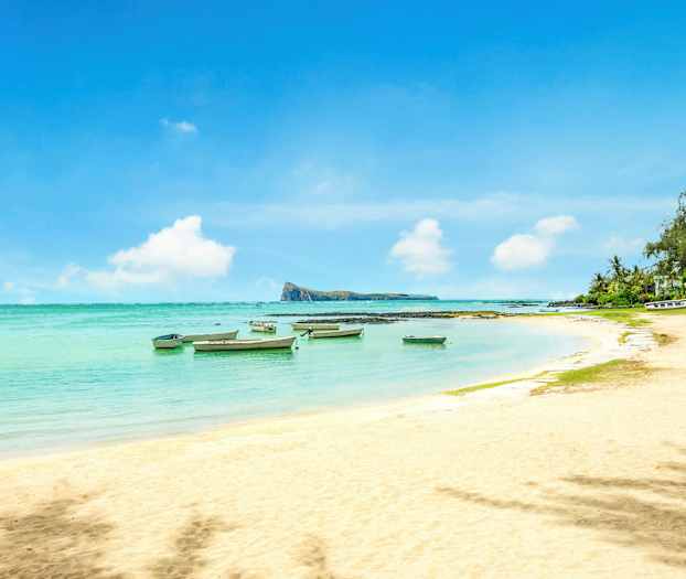 En sandstrand med småbåter i det klare vannet og vegetasjon ved kanten. (AI-generert tekst)