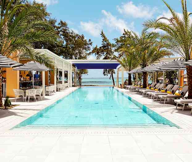 Svømmebasseng utendørs omgitt av solsenger, parasoller og palmer med hav og blå himmel i bakgrunnen. (AI-generert tekst)