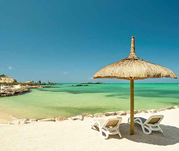 En sandstrand med to solsenger under en parasoll av strå, med turkist vann i bakgrunnen. (AI-generert tekst)