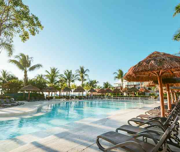 Et svømmebasseng omgitt av solsenger og palmer under en klar himmel. (AI-generert tekst)