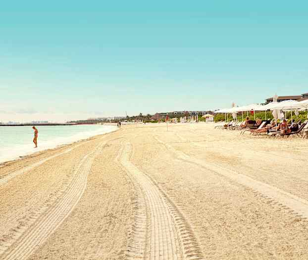 En sandstrand med parasoller og solsenger, en person står ved vannet. (AI-generert tekst)