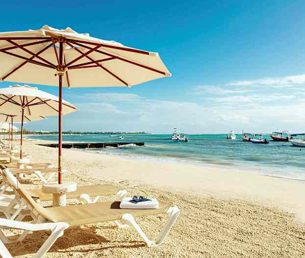En sandstrand med solsenger og parasoller. I bakgrunnen sees havet med båter. (AI-generert tekst)