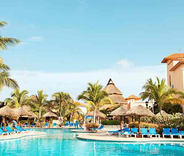 Bildet viser et utendørsbasseng omgitt av solsenger med blå puter og parasoller. I bakgrunnen ses palmer og bygninger med oransje tak. (AI-generert tekst)