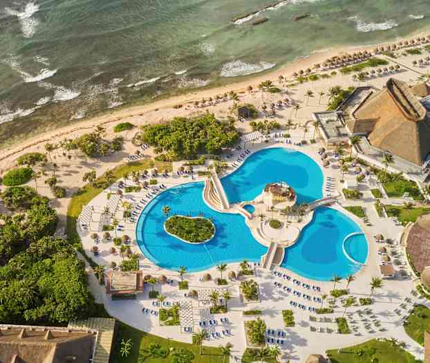 Luftfoto av et feriested ved sjøen med flere bassenger, grøntområder og bygninger med stråtak. (AI-generert tekst)