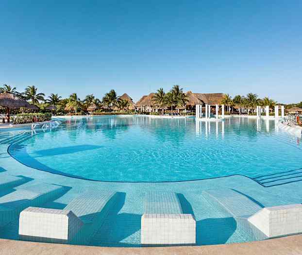 Et stort, klart blått svømmebasseng foran en rekke med palmer og stråhytter under en skyfri himmel. (AI-generert tekst)