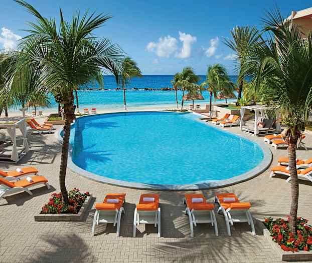 Et bassengområde med solsenger og parasoller, omgitt av palmer, med utsikt over havet i bakgrunnen. (AI-generert tekst)