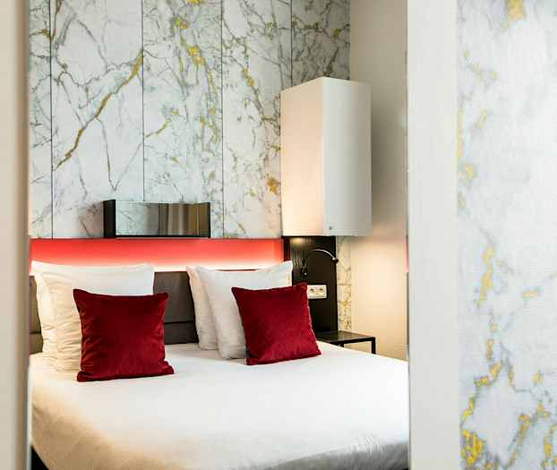 Et moderne hotellrom med en dobbeltseng, hvite og røde puter samt en bakgrunnsvegg med marmormønster. (AI-generert tekst)