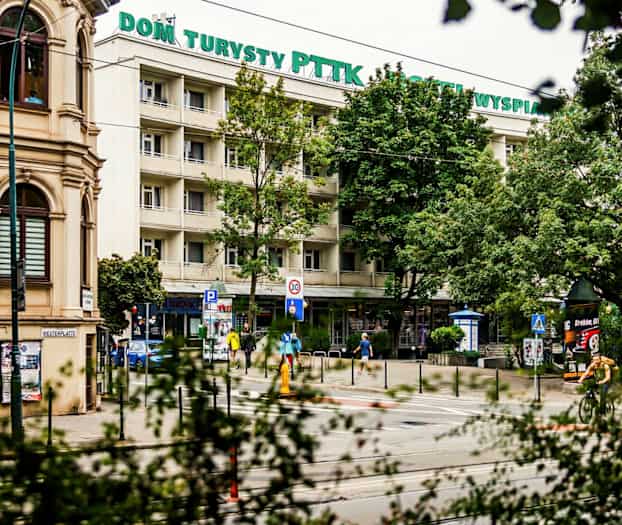 Et bilde av en bygning med skilt "Dom Turysty PTTK Hotel Wyspiański" omgitt av trær. På gaten foran sees mennesker, sykler og skilt. (AI-generert tekst)