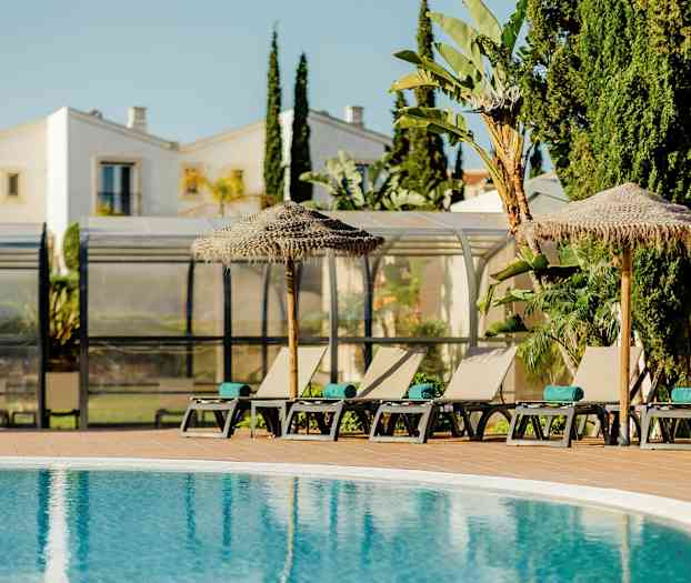 Solstoler og parasoller ved et basseng med bygninger og vegetasjon i bakgrunnen. (AI-generert tekst)