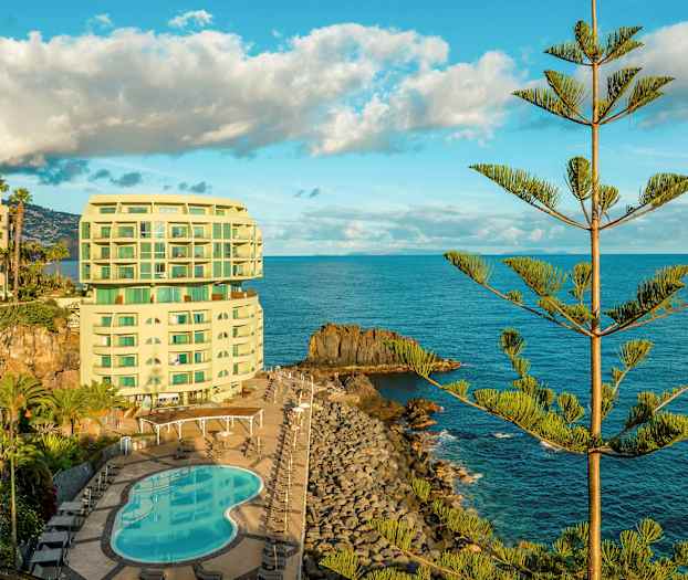 Et hotell ved kysten med et basseng og omgitt av hav og trær. (AI-generert tekst)