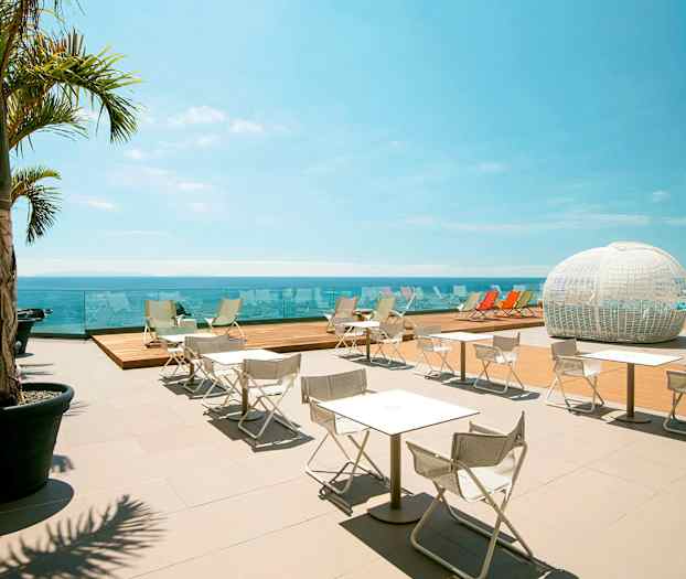 En utendørsterrasse med hvite stoler og bord, skyggelagt av palmer, utsikt over havet og en hvit, kuppelformet struktur i bakgrunnen. (AI-generert tekst)