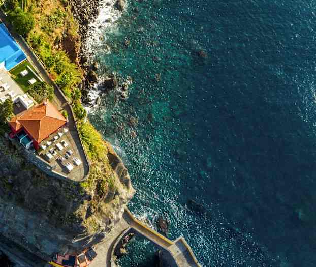 Dronebilde av en klippe med en bygning og basseng nær sjøen. (AI-generert tekst)