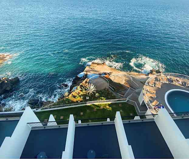 Luftfoto av et anlegg ved kysten med blått hav, klipper, basseng og soldekk. (AI-generert tekst)